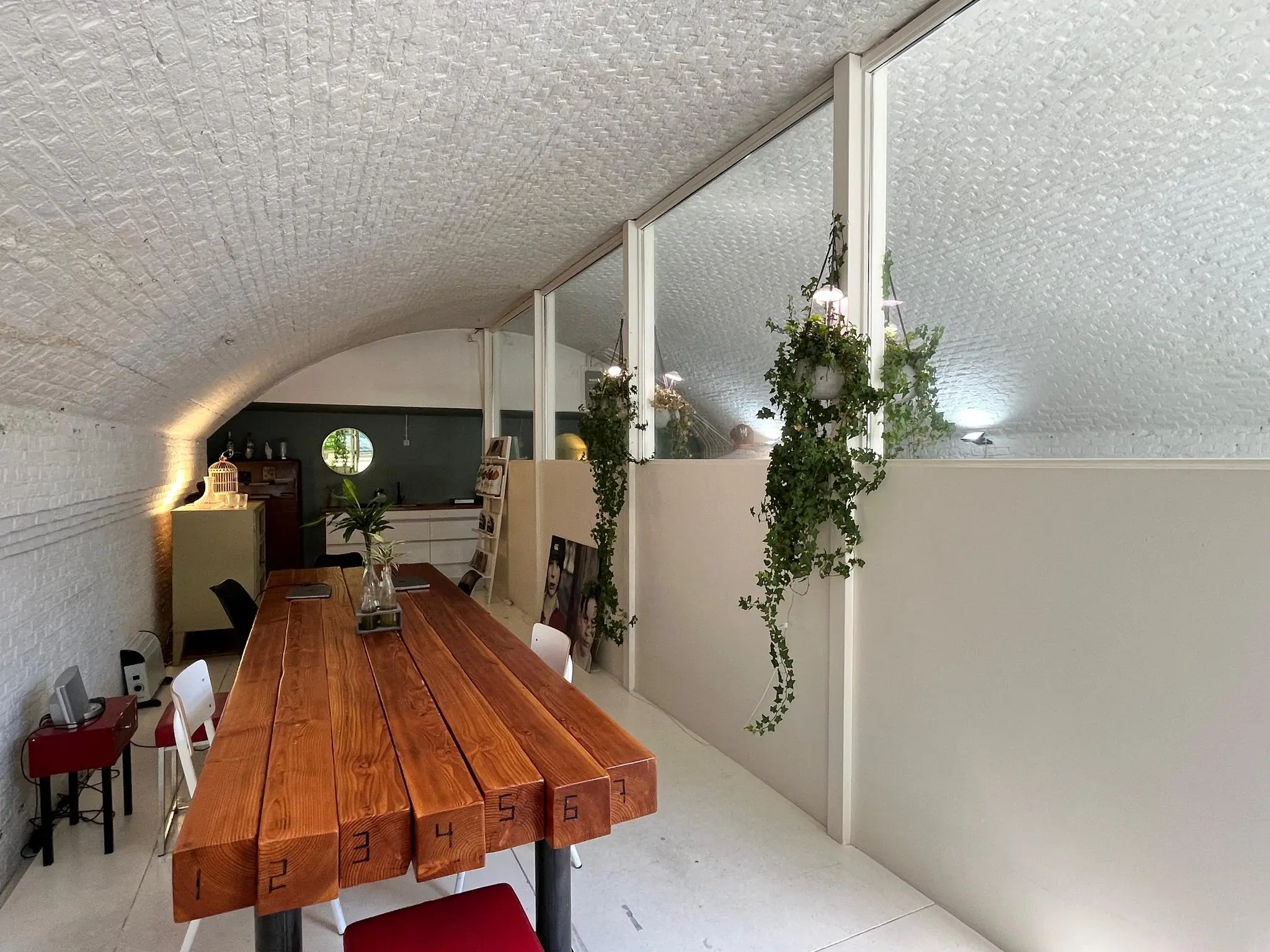 Langwerpige ruimte met gewelfd plafond, houten vergadertafel met genummerde balken, glazen wand en hangende planten in interieur "Tussen de Bogen".